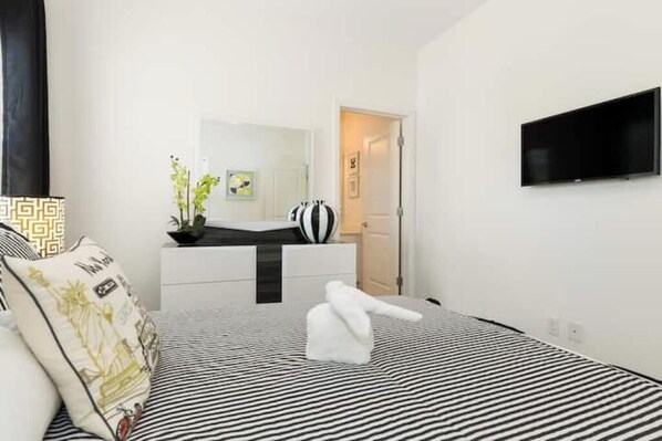 Una camera d'albergo con un letto, una TV a muro, un comodino e una porta che dà su un'altra stanza.