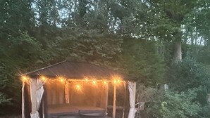 A gazebo with string lights, a hot tub, and steps leading to it, surrounded by a grassy area and trees.