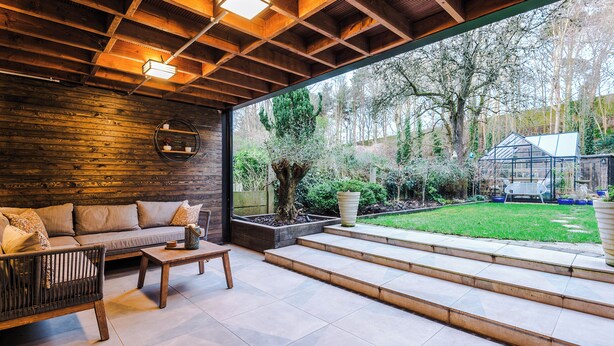 A cozy patio area with a wooden ceiling, a sofa, a coffee table, and a view of a garden with a tree and a greenhouse.