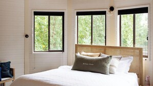A bedroom with a wooden ceiling, a large bed with a headboard, two windows, and a bench. - Beltana Farm (Pialligo)