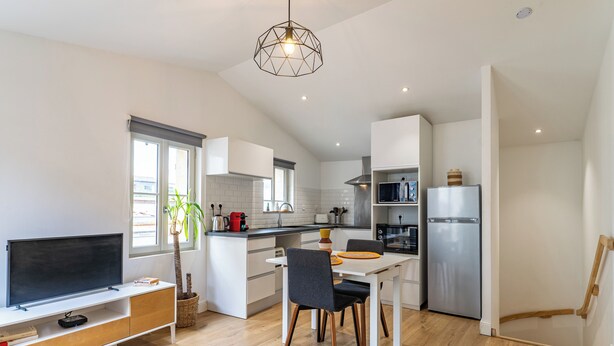 A modern kitchen with white cabinets, a built-in oven, and a microwave. There's a dining area with a table and chairs, and a TV on a stand. - Duplex flat for 2 people with terrace and parking (Bordeaux)