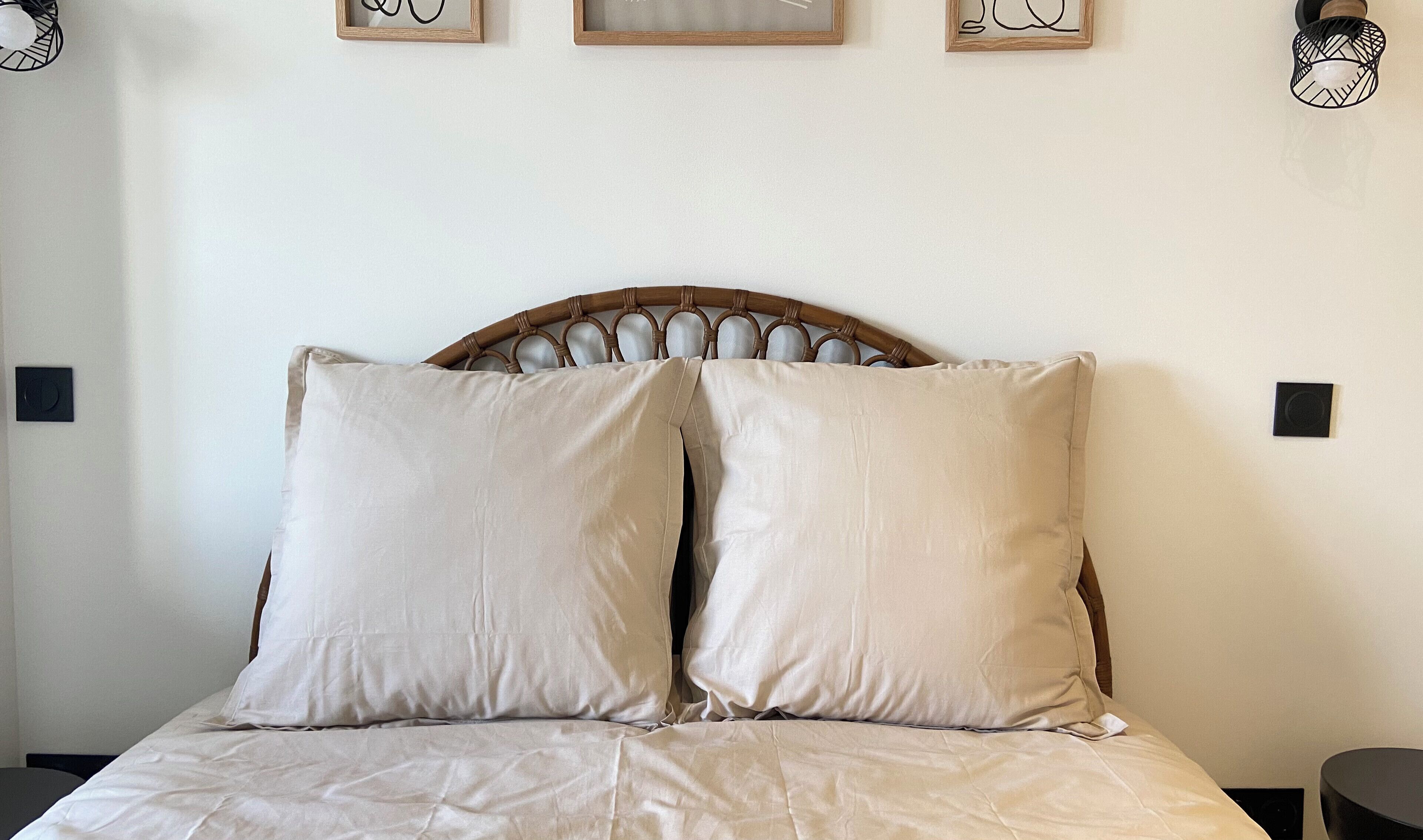 Uma cama com cabeceira de madeira, duas almofadas e uma parede lisa com um quadro e um ponto de luz.