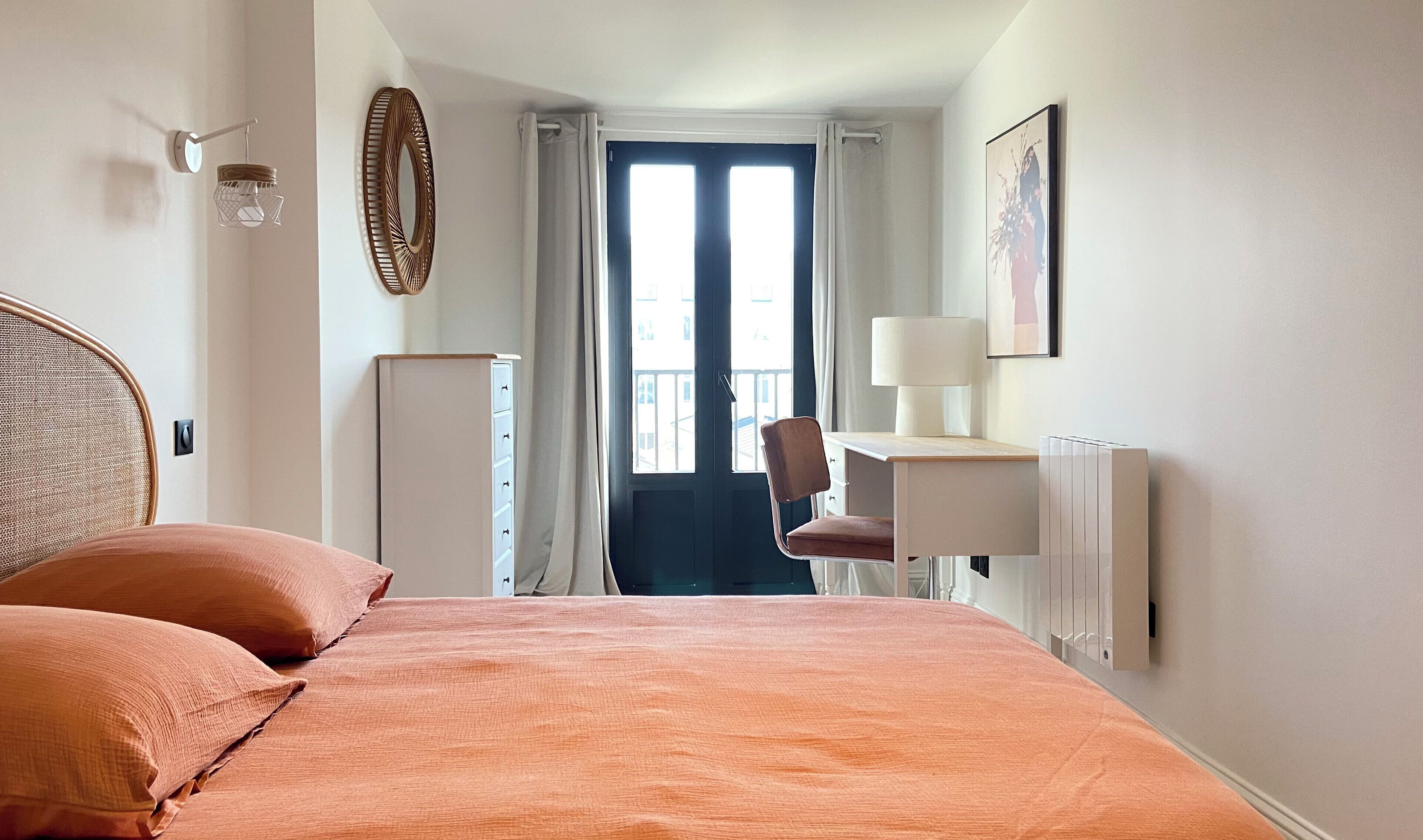 Ein Schlafzimmer mit einem Bett, einem Sessel, einem Schreibtisch, einem Heizkörper, einem Spiegel und einem Fenster mit Vorhängen.