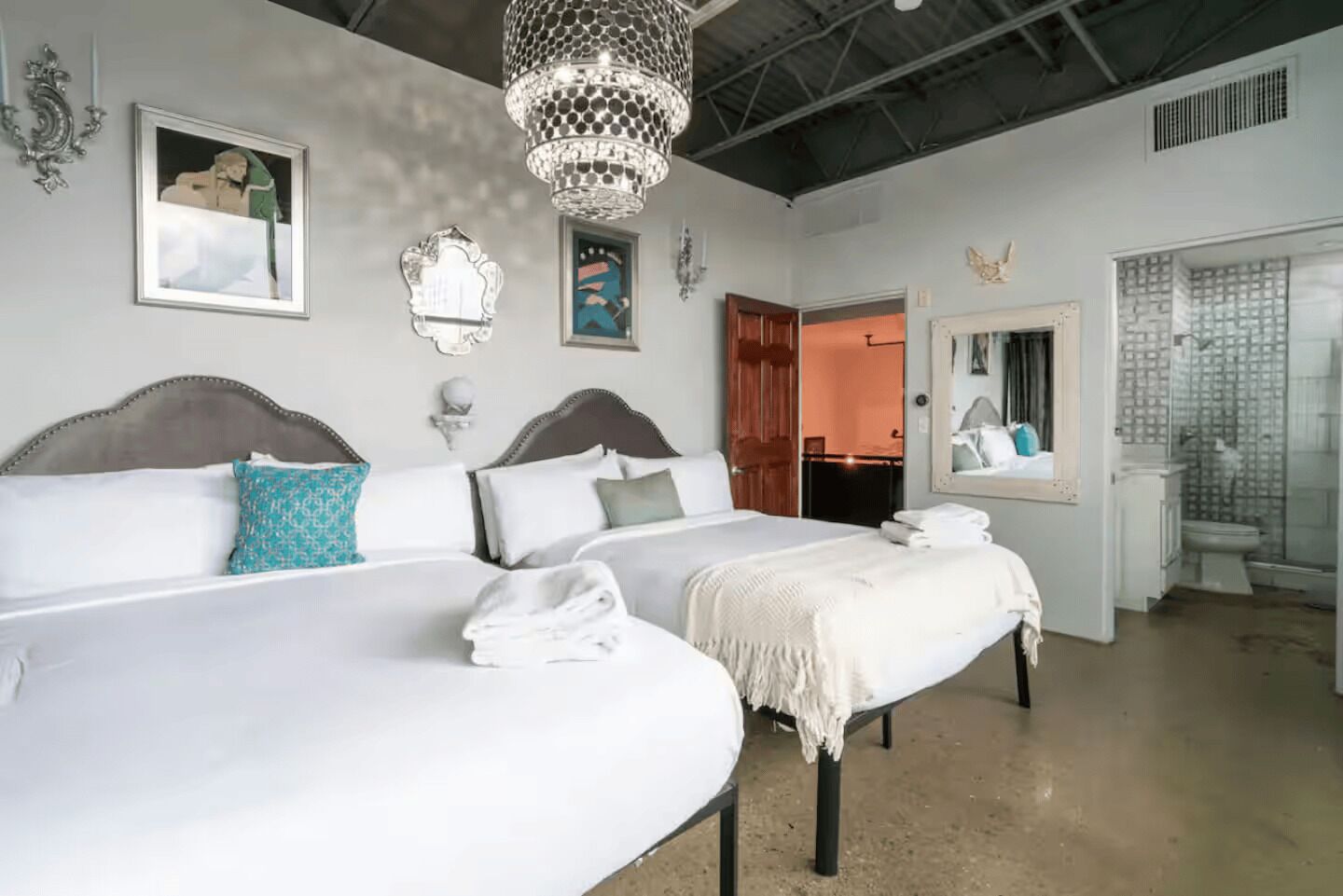 A hotel room with two beds, a chandelier, artwork on the walls, and a bathroom with a shower and toilet.