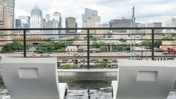 Vista desde un balcón con dos sillas blancas que dan a un paisaje urbano con edificios modernos y una calle concurrida abajo.