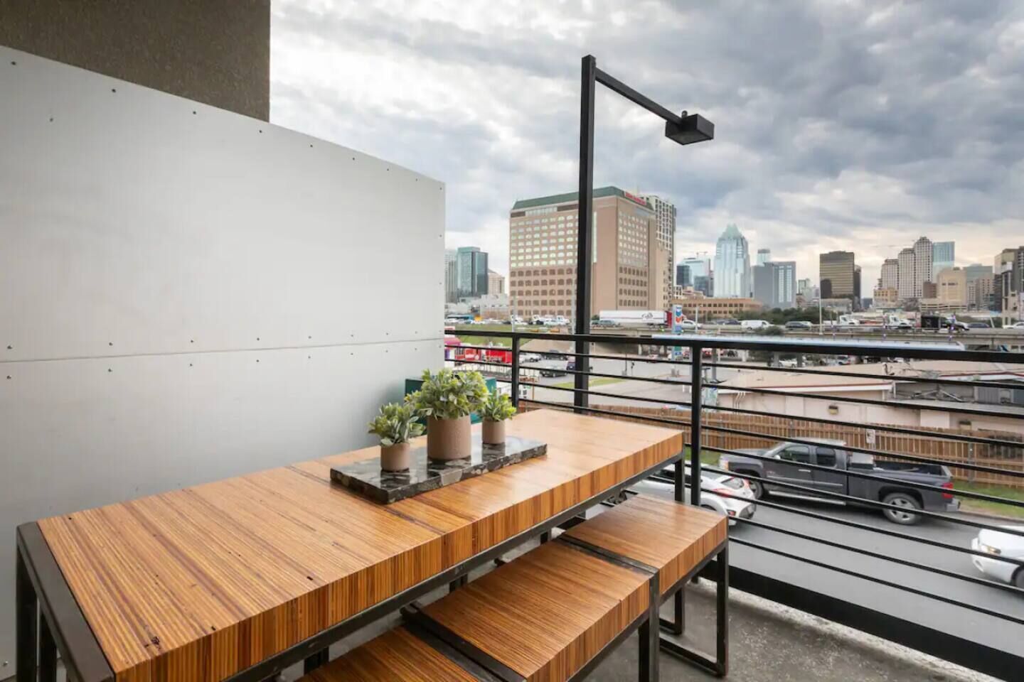A balcony with a wooden table and chairs, potted plants, and a city view.