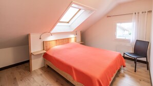 A bedroom with a sloped ceiling, a bed with an orange bedspread, a chair, and a window with curtains. - MAISON MARMILLONS - CANOPEE (AIX LES BAINS)