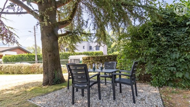 An outdoor seating area with a table and chairs, surrounded by gravel and grass, with a tree and a hedge in the background. - MAISON MARMILLONS - CANOPEE (AIX LES BAINS)