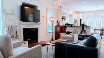 A living room with a fireplace, a flat-screen TV, and a dining area with a kitchen in the background.
