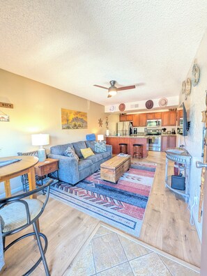 A living room with a sofa, coffee table, and a dining area with chairs. There is a kitchen in the background with wooden cabinets and appliances. - ~Red Rock Sanctuary w/Views~Pool~BBQ~Cozy~Clean~ (Sedona)