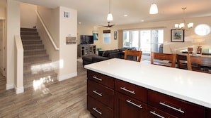 A modern kitchen with a white countertop, wooden cabinets, and a built-in oven. There is a staircase leading to another level and a dining area with a table and chairs.