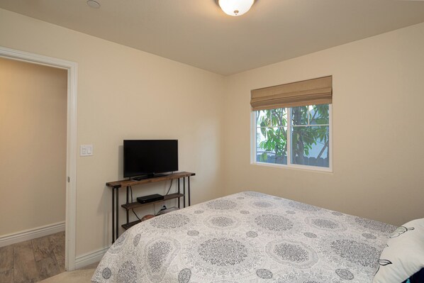 Un dormitorio con cama, televisión sobre un mueble, ventana con vistas a árboles y una puerta.