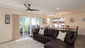 A living room with a brown leather sofa, a ceiling fan, a dining area with a wooden table and chairs, and a sliding glass door leading to an outdoor patio.