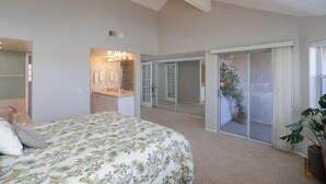 Een slaapkamer met een bed, een badkamer met een spiegel en verlichting, een glazen schuifdeur naar een balkon en een potplant.