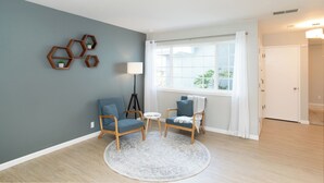 Una habitación con dos sillas, una mesita redonda pequeña, una lámpara y un ventanal con cortinas.