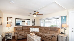A living room with a brown leather sectional sofa, a wooden coffee table, two side tables with lamps, a ceiling fan, and a window with blinds.