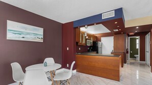 A modern kitchen with a central island, white chairs, and a painting of a beach scene. - Bay Harbor Sunset Apt 2A (Bay Harbor Islands)
