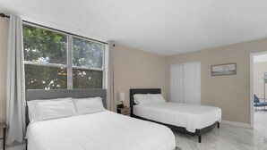A bedroom with two beds, a window with curtains, a bedside table with a lamp, and a door leading to another room. - Bay Harbor Sunset Apt 2A (Bay Harbor Islands)