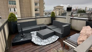 Eine Dachterrasse mit Holzdeck, grauer Gartenmöbeln und einem gemusterten Teppich.