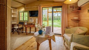 Ein gemütliches Holzhaus-Interieur mit Fernseher, einem Tisch mit einer Vase, Korbstühlen und einer großen Schiebeglastür zum Garten.