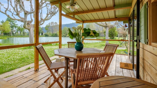 Eine Holzterrasse mit Tisch und Stühlen, einer Hängelampe und Blick auf einen See und Bäume.
