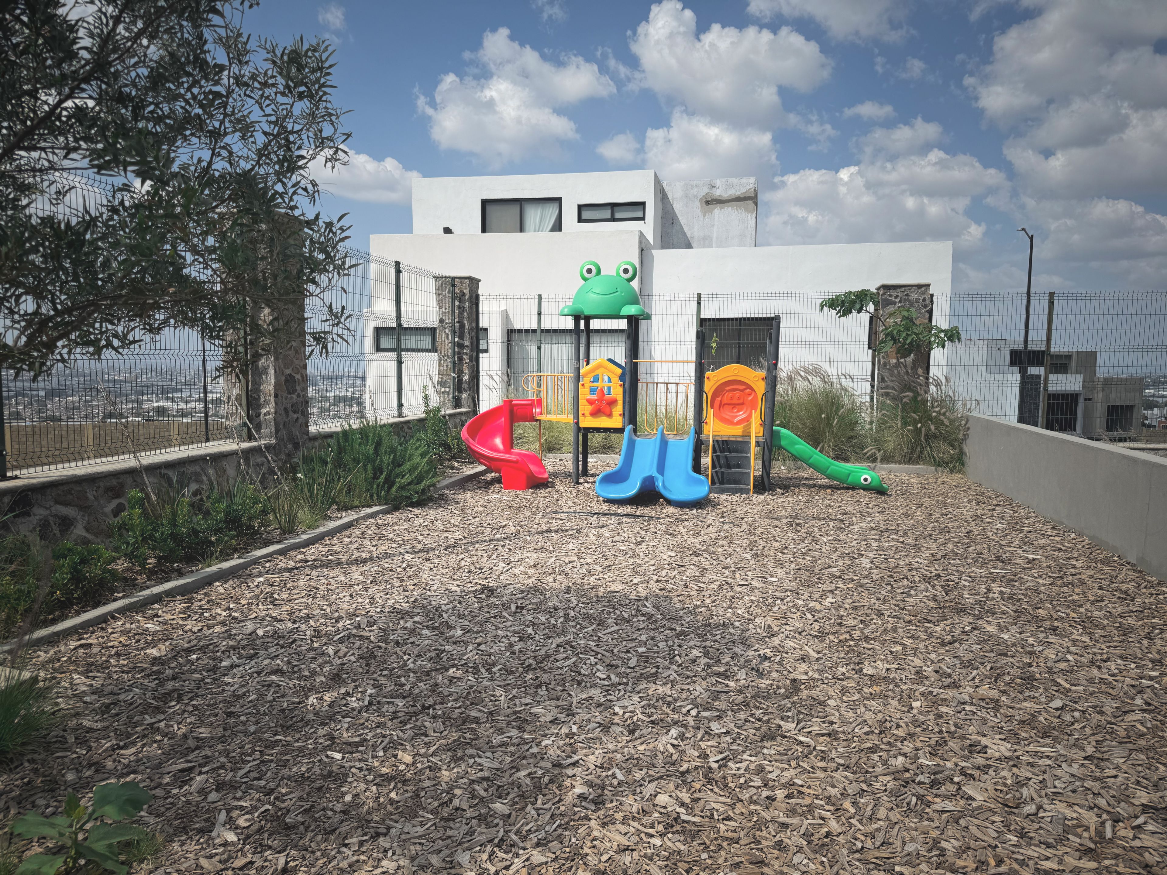 Un parque infantil con toboganes y estructuras para escalar, rodeado por una cerca y un edificio al fondo.