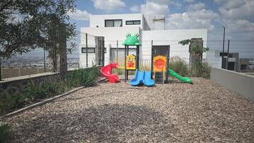 Un parque infantil con toboganes y estructuras para escalar, rodeado por una cerca y un edificio al fondo.