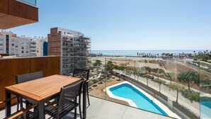 A balcony with a wooden table and chairs, a swimming pool, and a view of the beach and ocean.