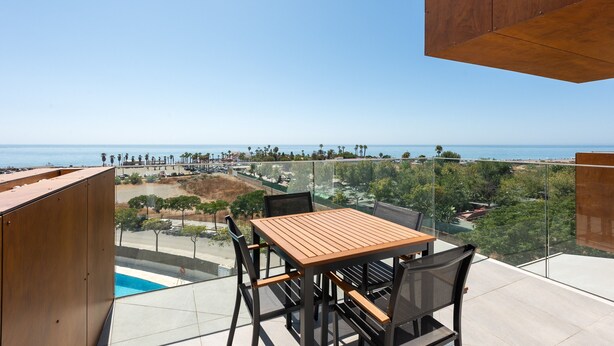 A balcony with a wooden table and chairs, overlooking a coastal view with a pool and greenery.