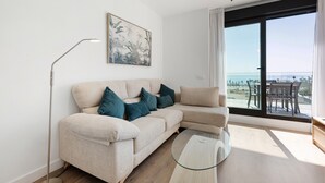 A modern living room with a beige sofa, a glass coffee table, and a large window with a view of the sea.