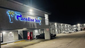 Un motel con un letrero de neón que dice "Paradise Inn" y un gráfico de palmera. Hay coches estacionados al frente, una máquina expendedora de Coca-Cola y una escalera que lleva a la entrada.