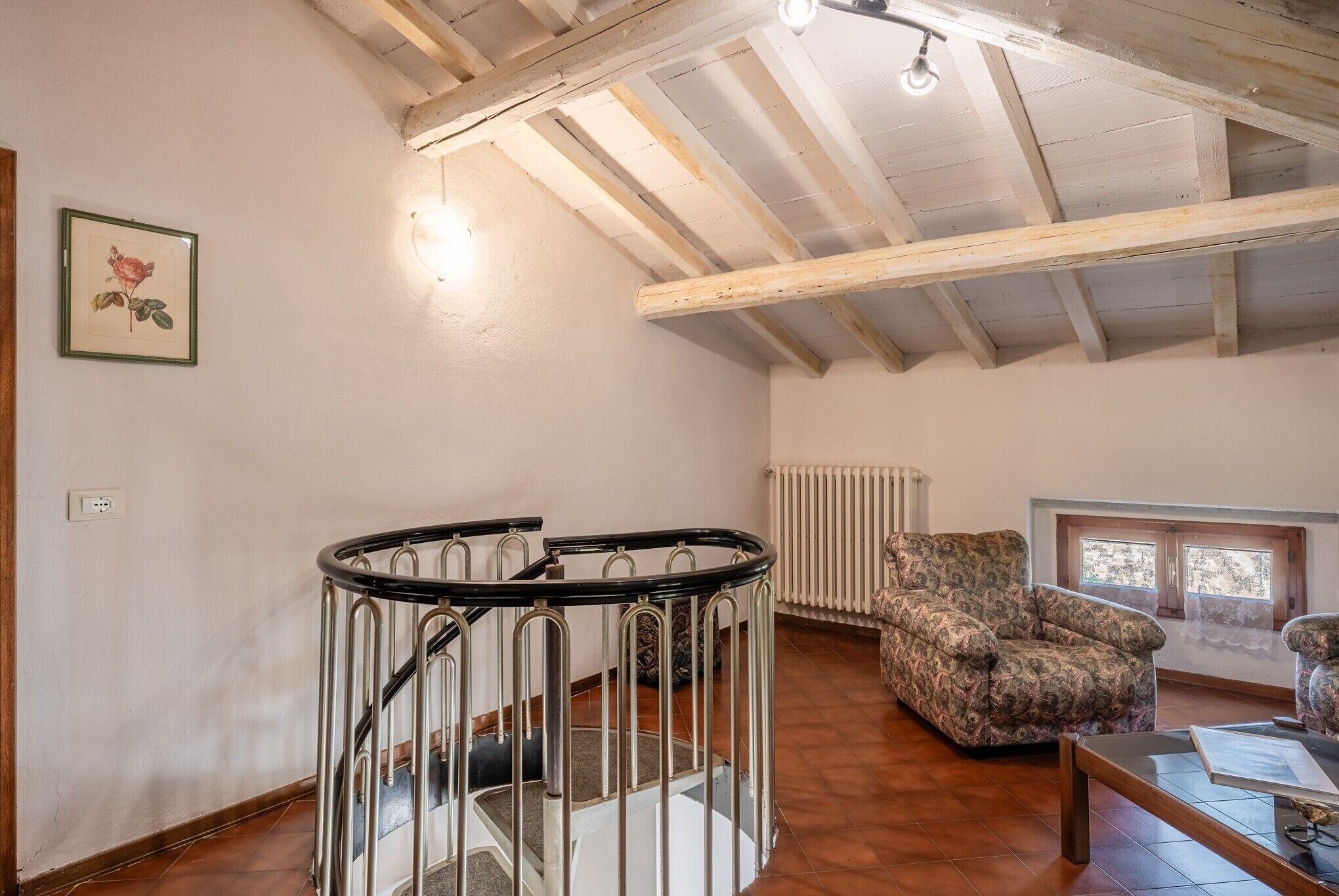 A room with a wooden ceiling, a staircase with metal railings, a radiator, a patterned sofa, and a table with papers.