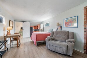 Un dormitorio con una cama, un sillón gris, una mesa de comedor con sillas y una cocina al fondo.