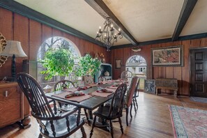 Ein Esszimmer mit einem langen Tisch, Stühlen und einer Chandelier. Es gibt Fenster mit Pflanzen und eine Tür im Hintergrund.