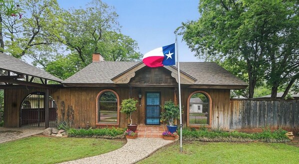 A rustic house with a Texas flag, a wooden porch, and a brick chimney.