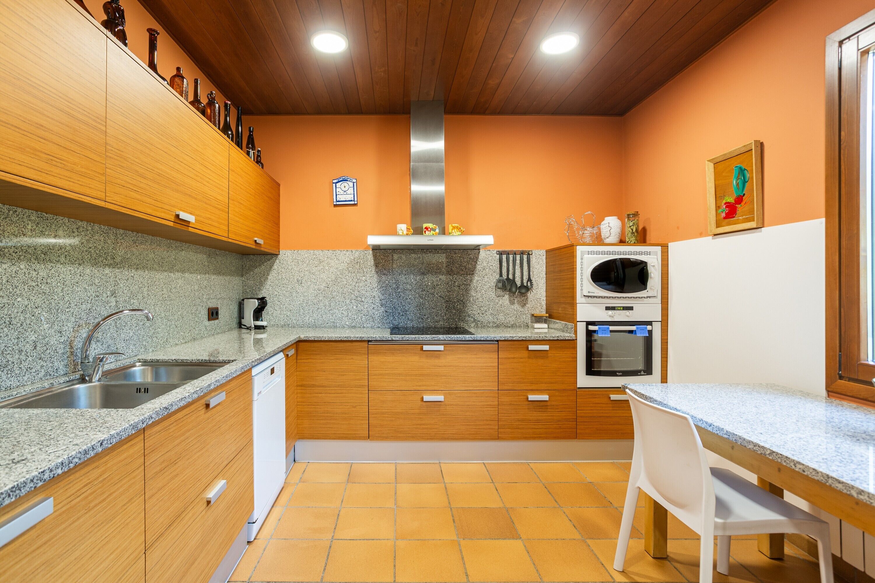 Una cocina con encimeras de granito, armarios de madera, campana extractora de acero inoxidable, horno empotrado y una silla blanca.
