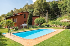 Una piscina con borde de baldosas, rodeada por un patio cercado, una casa de fachada roja y una colina con árboles.