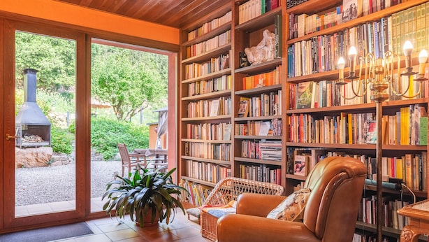 Uma sala de leitura acolhedora, com lareira, estante de livros, poltrona de vime e um vaso de plantas.