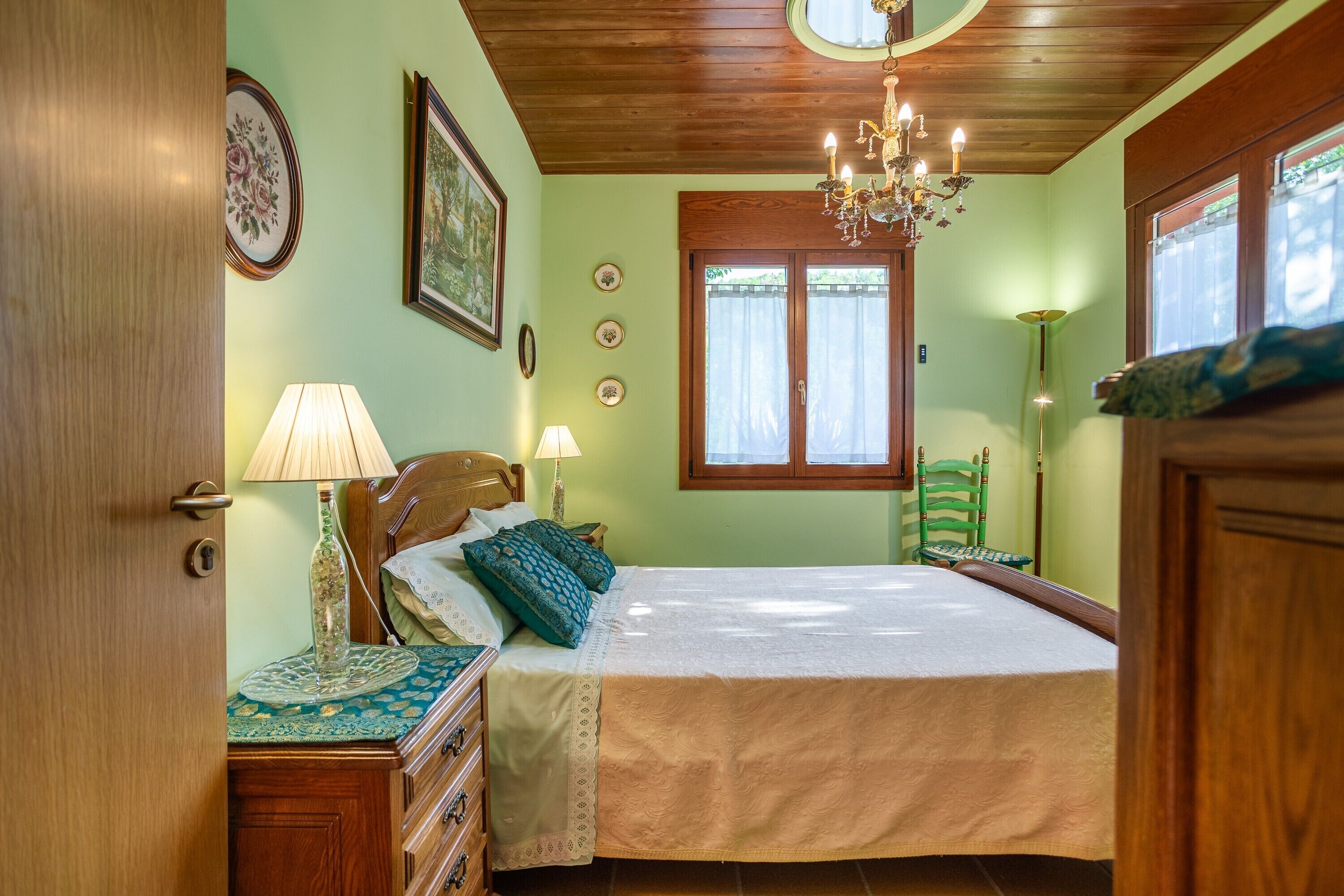 Un dormitorio con techo de madera, una cama con colcha blanca, una mesita de noche con lámpara, un sillón verde y una ventana con cortinas.