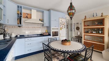 Une cuisine avec des meubles de rangement bleus, une table de salle à manger ronde entourée de chaises, un luminaire suspendu et un meuble en bois doté de portes en verre.
