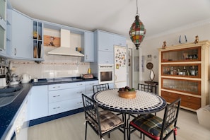 Eine Küche mit blauen Schränken, einem runden Esstisch mit Stühlen, einer Pendelleuchte und einem Holzschrank mit Glastüren.