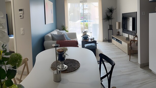 Un salon moderne avec une table à manger blanche, un canapé, un téléviseur et une grande fenêtre avec des rideaux.