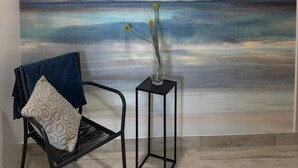 Une pièce ornée d’une grande fresque murale représentant une mer et un ciel paisibles, d’une chaise en métal noir avec un coussin, et d’une petite table sur laquelle est posé un vase.