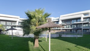 A modern building with a grassy courtyard, palm trees, and thatched umbrellas. - Apartment 'Palacio De Pilas 22' with Shared Pool, Wi-Fi and Air Conditioning (Zahara de los Atunes)