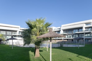 A modern building with a grassy courtyard, palm trees, and thatched umbrellas.