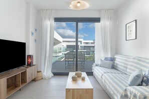 A modern living room with a flat-screen TV, a sofa, a coffee table, and a balcony with a view of buildings.