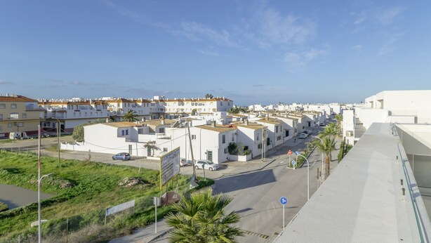 A coastal residential area with white buildings, a clear sky, and a road with cars.