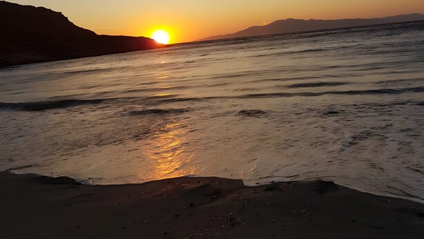 Une plage où le soleil se couche sur l’eau, avec un ciel dégagé et des montagnes à l’horizon.