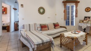 Un salon chaleureux avec un canapé, une table à manger recouverte d’une nappe en dentelle, un fauteuil en osier et une fenêtre avec des rideaux.