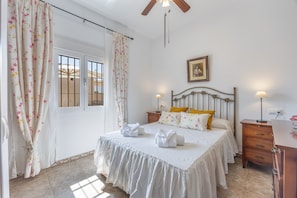 A bedroom with a bed, two bedside tables, a ceiling fan, a window with curtains, and a wall-mounted picture.